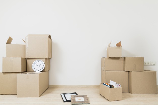 stacked-moving-cardboard-boxes-with-clock-picture-frame-against-white-wall_23-2148060008